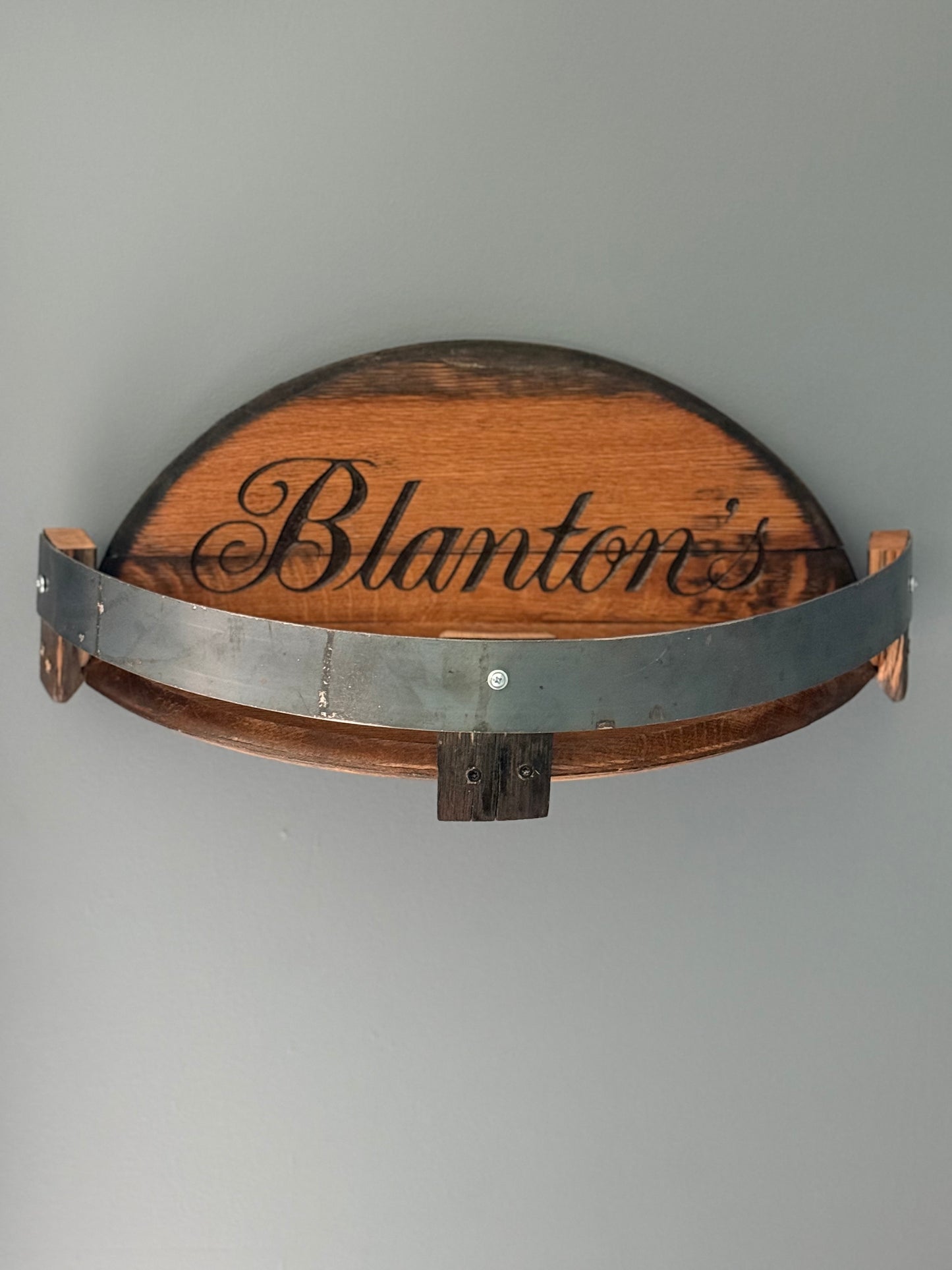 Bourbon barrel lids split to create a shelf bordered by small pieces of stave with metal barrel band. Lid engraved with "Blanton's".