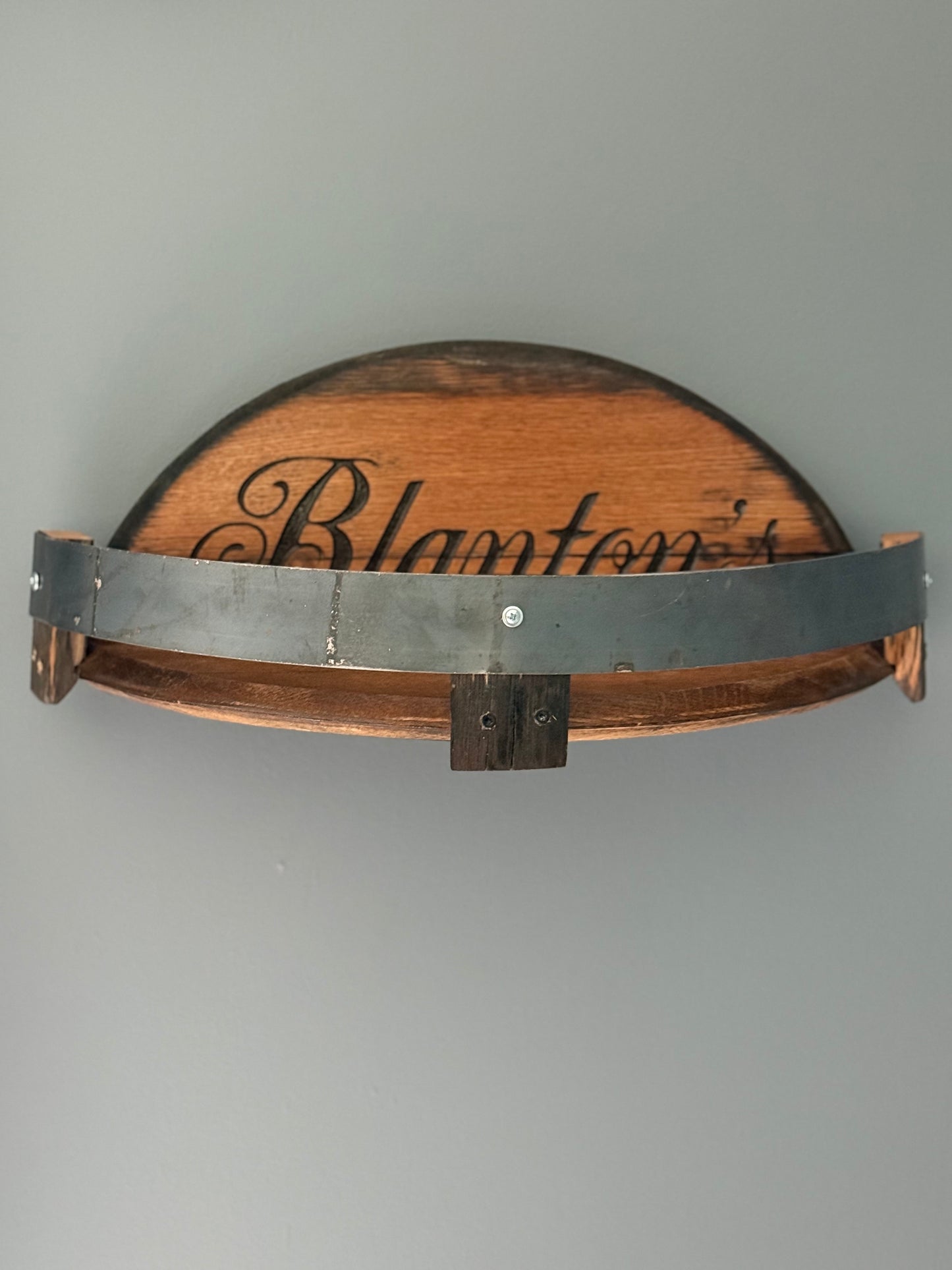 Bourbon barrel lids split to create a shelf bordered by small pieces of stave with metal barrel band. Lid engraved with "Blanton's".