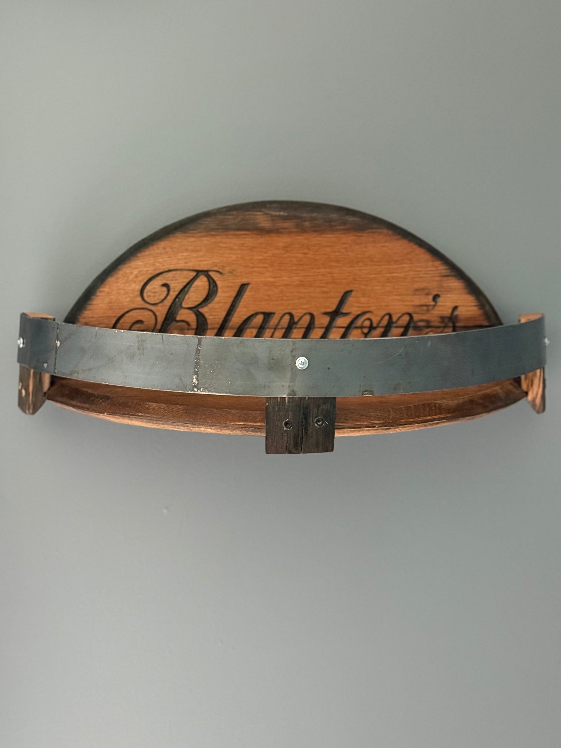 Bourbon barrel lids split to create a shelf bordered by small pieces of stave with metal barrel band. Lid engraved with "Blanton's".